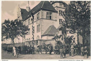 Black and white photo of a group of people standing in front of a building with windows, trees, and a sky in the background, with text at the bottom reading "Einzug in die neue Kasemme am 1 oktober 1909".