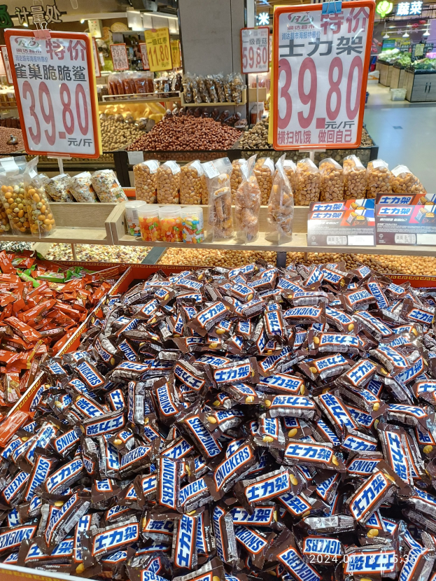 Ein Vitrinenschrank voller verschiedener Süßigkeiten, darunter Snickers-Riegel, in einem Geschäft mit Lebensmittelartikeln auf Regalen und Deckenlampen im Hintergrund.