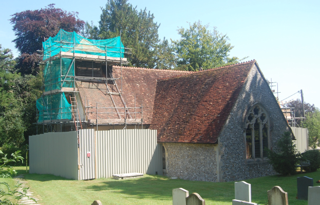 Kirche mit einer grünen Plane auf dem Dach, umgeben von einem Metallzaun, Bäumen, Pflanzen, Gras, einem Pfad, Pfosten und einem bewölkten Himmel.