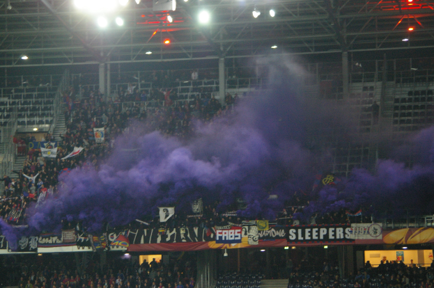 Große Menschenmenge in einem Stadion mit violettem Rauch, Schildern mit Text, Stühlen, Treppen, Geländern und Deckenlampen.