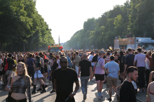 Eine große Menschenmenge, die eine von Bäumen gesäumte Straße entlanggeht, mit einem Turm im Hintergrund, Fahrzeugen mit Menschen auf der rechten Seite, wahrscheinlich beim Christopher Street Day Festival unter einem klaren blauen Himmel.