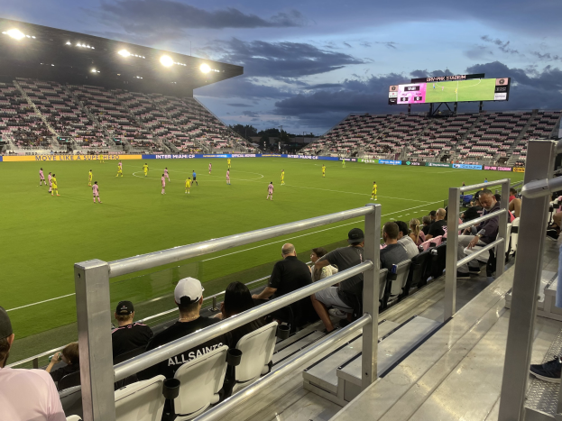 A soccer game between Orlando City SC and Philadelphia Union being played in a stadium with spectators in the stands, scoreboard visible, surrounded by trees under a cloudy sky.