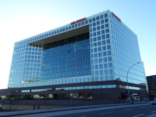 Large building with many windows, identified as the Deutsche Bank headquarters, surrounded by street poles, street lights, motor vehicles, pedestrians, trees, and a cloudy sky.