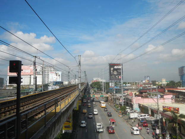 A bustling city street with heavy traffic, a railway track on the left, electric poles with wires, a traffic signal, buildings, trees, hoardings, and a cloudy sky.