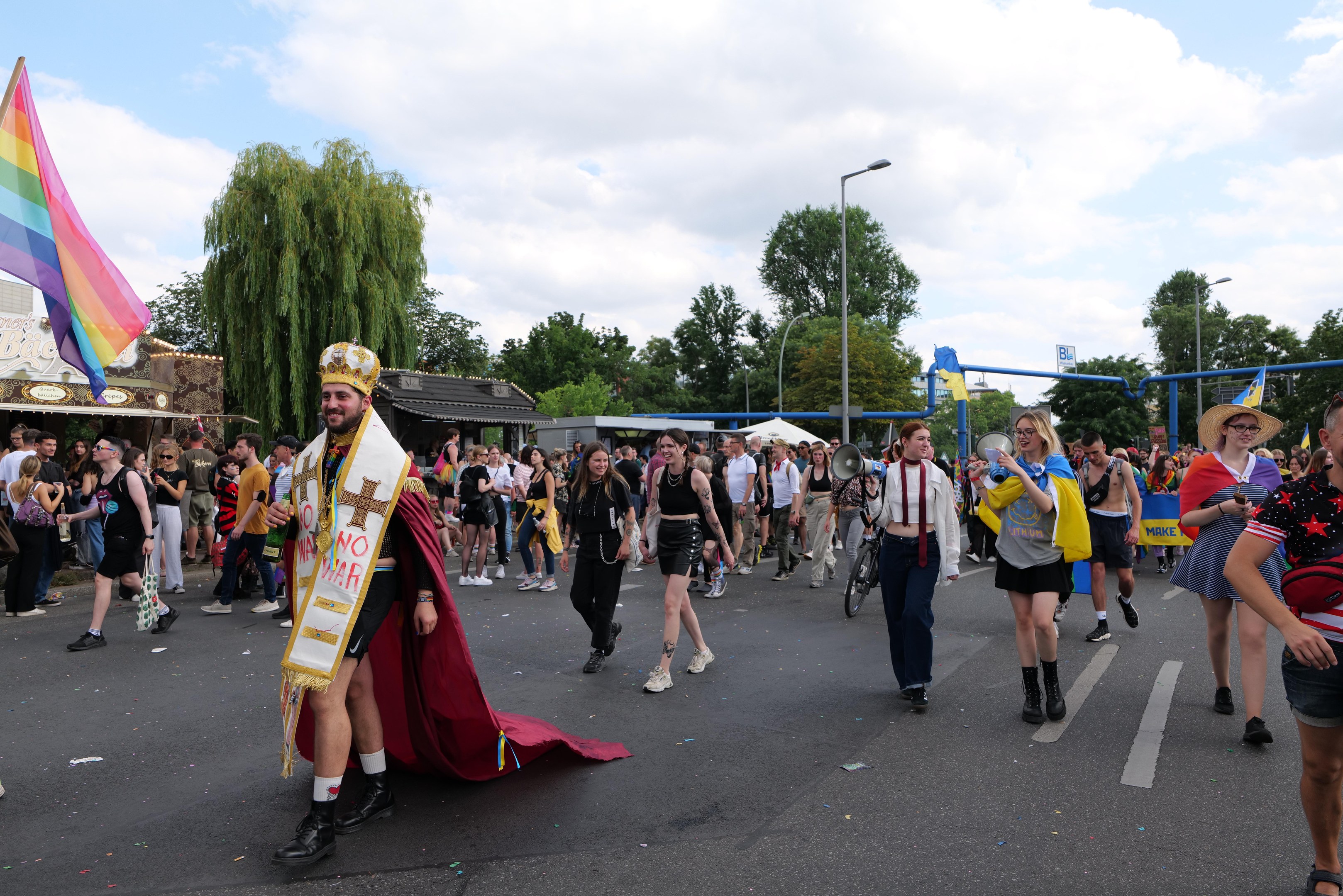 Eine Gruppe von Menschen, die bei der Gay Pride Parade 2018 marschieren und eine Regenbogenflagge sowie Musikinstrumente tragen, mit Laternenmasten, Bäumen und Hütten im Hintergrund.