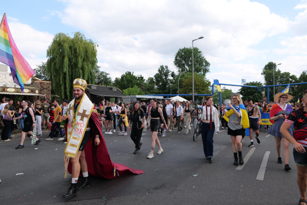Eine Gruppe von Menschen, die bei der Gay Pride Parade 2018 marschieren und eine Regenbogenflagge sowie Musikinstrumente tragen, mit Laternenmasten, Bäumen und Hütten im Hintergrund.