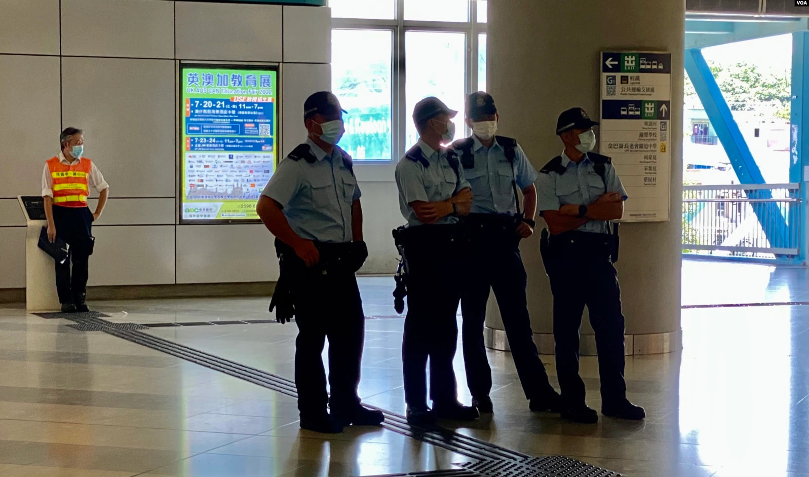 Eine Gruppe von Polizisten in Masken steht vor einem Gebäude mit Glastüren, Schildern mit Text, einer Säule und einem Geländer, mit Bäumen und Fahrzeugen im Hintergrund.