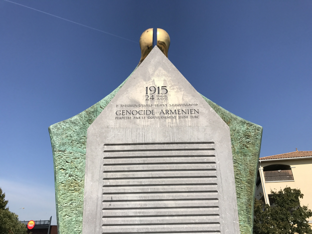 Ein hoher Denkmal mit einer Uhr an seiner Spitze steht in einem Park, umgeben von Bäumen und Gebäuden unter einem klaren Himmel, mit eingravierter Text, der den 24. Jahrestag des Völkermords in Armenien gedenkt.