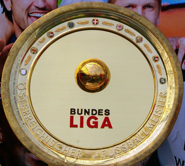 Ein Mann hält einen goldenen Schild mit der Aufschrift "Bundesliga" hoch, im Hintergrund eine Fahne mit drei Personen