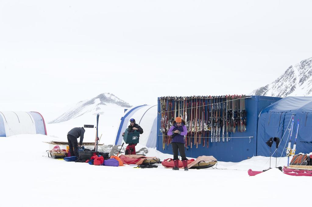 Drei Personen stehen auf einer schneebedeckten Landschaft mit Taschen drumherum, Zelte mit Skiern dahinter und schneebedeckte Hügel im Hintergrund unter einem klaren Himmel.