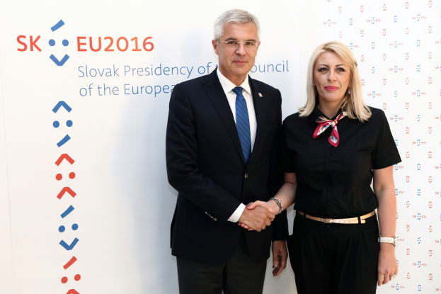 Zwei Personen, ein Mann in einem schwarzen Blazer und weißem Hemd mit blauer Krawatte und eine Frau mit Brille, stehen lächelnd vor einer weißen Tafel mit der Aufschrift "sk eu 2016", identifiziert als Präsident des Europäischen Rates.