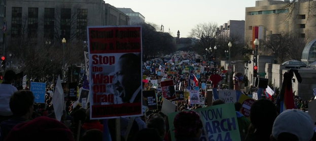 People protesting on a road holding posters, with buildings, trees, light poles, and the sky in the background.