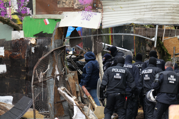 Group of police officers in black uniforms and helmets standing in front of a destroyed building surrounded by a metal fence, with scattered debris.