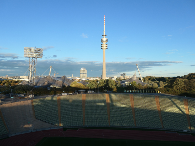 Olympiastadion in Berlin, Deutschland, mit dem Fernsehturm (Fernsehturm) im Hintergrund, umgeben von Bäumen, Gebäuden und beleuchteten Bereichen unter einem bewölkten Himmel.
