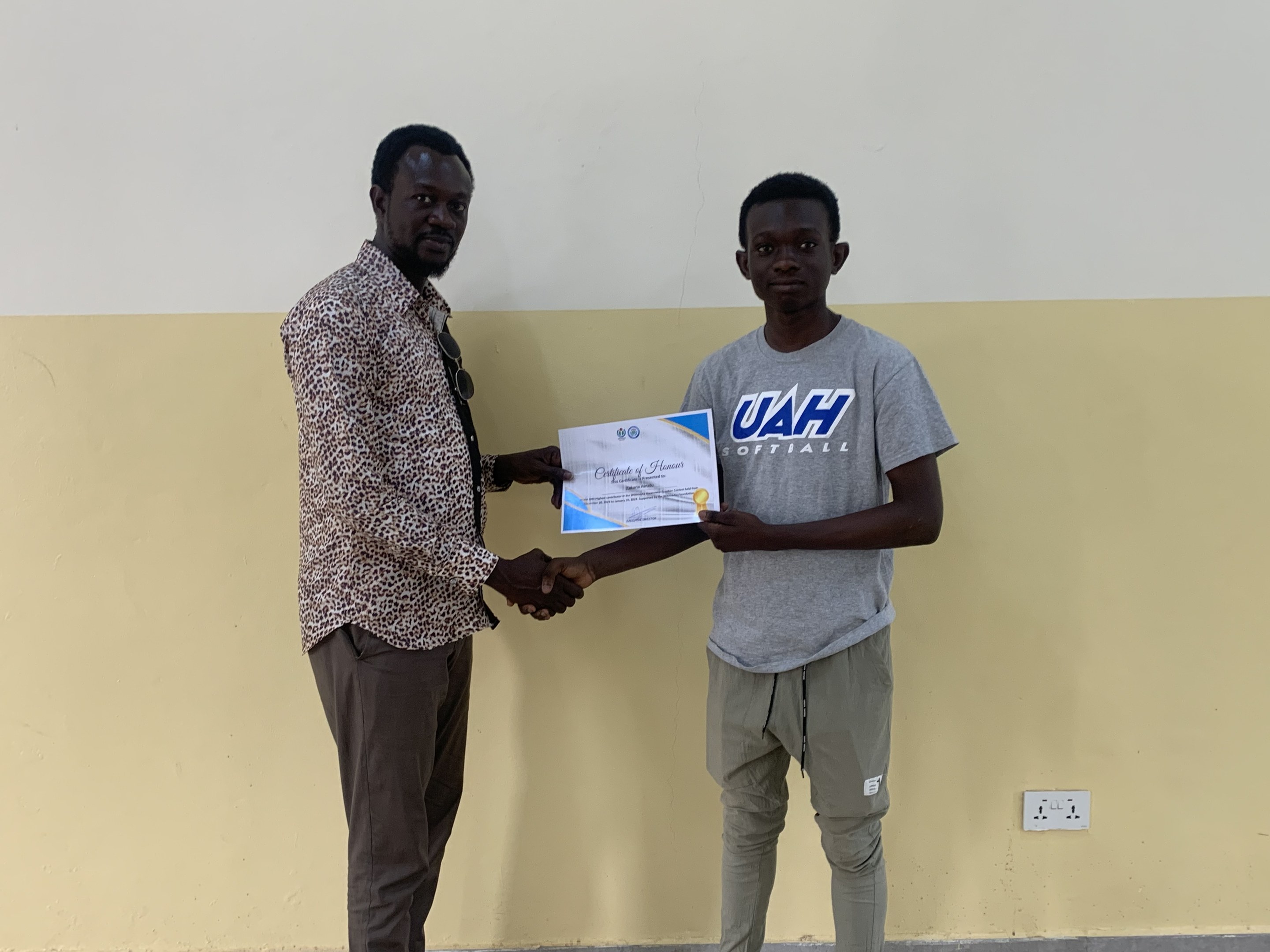 Zwei Männer, die vor einer gelben Wand die Hände schütteln, wobei der Mann links ein Zertifikat hält, nachdem er ein Stipendium von der Universität Ghana erhalten hat.
