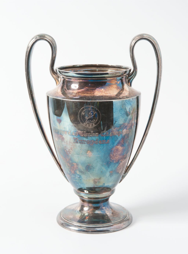 Der UEFA-Champions-League-Pokal, eine stählerne Tasse mit zwei Griffen und einem blauen und silbernen Farbschema, die mit "UEFA Champions League" beschriftet ist, vor einem weißen Hintergrund dargestellt.