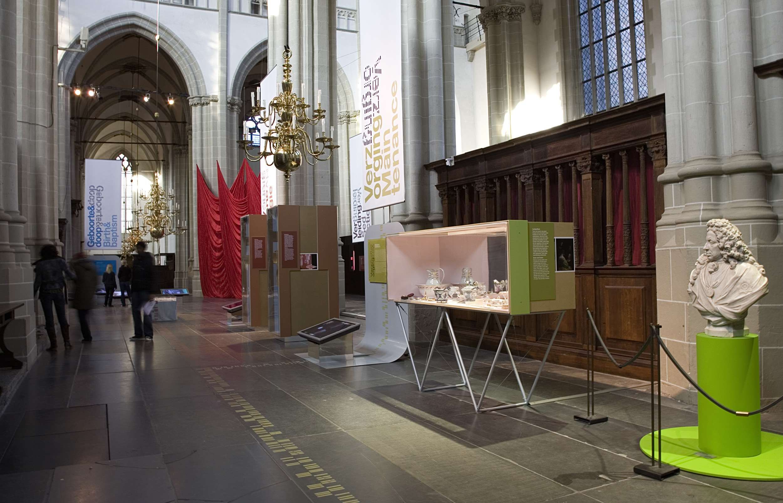 Ein großer Museumsraum mit verschiedenen Artefakten, darunter eine Statue auf einem Sockel, Absperrbänder, Textbanner, Säulen und Kronleuchter, sowie einige Personen.