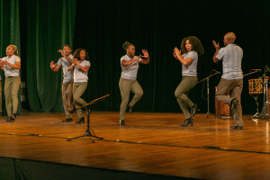 Eine Gruppe von Menschen auf einer Bühne mit Mikrofonen, Musikinstrumenten und einem Vorhang im Hintergrund, die Hände klappend in Feierlaune, scheinbar ein Tanzteam.