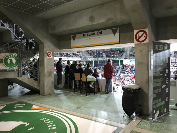 Eine Gruppe von Menschen steht vor einer sitzenden Menge in einem Stadion, mit Schildern, die "Boston Celtics vs New York Jets" anzeigen, einem Müllcontainer, einer Tafel mit Text, einer Treppe mit Geländern und Deckenleuchten.