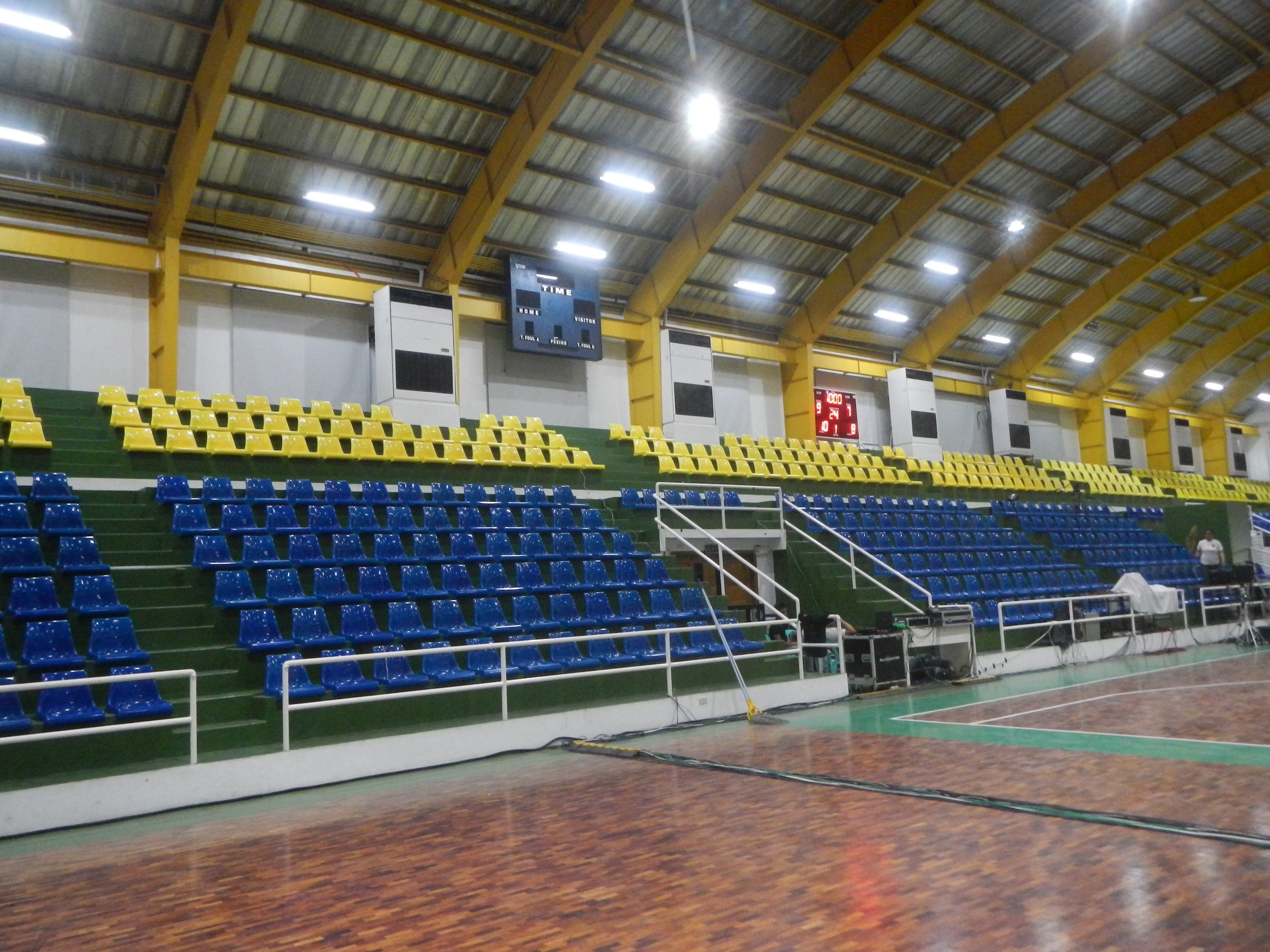 Großer Hallensportplatz mit Holzboden, blauen und gelben Sitzplätzen, Geländern, Treppen mit Geländern, wandmontierten Bildschirmen, Deckenleuchten und einigen Personen.