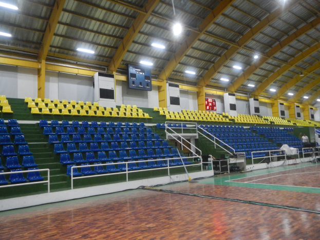 Großer Hallensportplatz mit Holzboden, blauen und gelben Sitzplätzen, Geländern, Treppen mit Geländern, wandmontierten Bildschirmen, Deckenleuchten und einigen Personen.