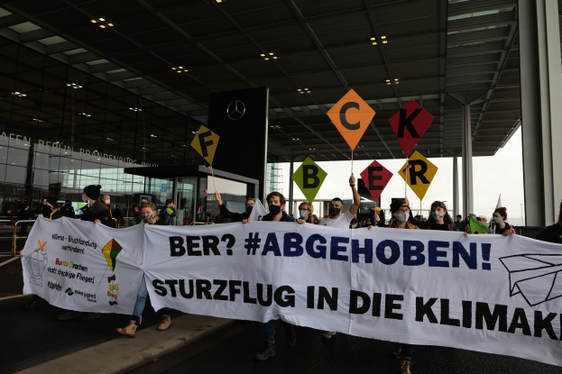 People wearing masks hold a banner reading "Ber? Abgehoben Sturzflug in die Klimakt" in front of a glass-walled building with illuminated pillars.
