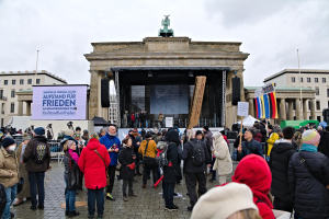 Eine Menschenmenge steht vor einem Gebäude mit einer Bühne, auf der Redner und ein Bildschirm zu sehen sind, sowie rechts davon Fahnen und Transparente, was auf eine Protestveranstaltung in Berlin hindeutet.
