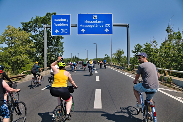 Eine Gruppe von Radfahrern in Helmen, die eine Straße mit einem Geländer und Bäumen auf beiden Seiten hinunterfahren, Lichtmasten im Hintergrund und ein klarer blauer Himmel, mit einem Schild, das eine Radtour in Hamburg anzeigt.