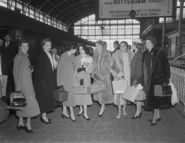 Schwarz-weißes Bild einer Gruppe von Frauen an einem Bahnhof, jede hält eine Tasche, mit einem Zug links und Geländern und einer Tafel rechts.