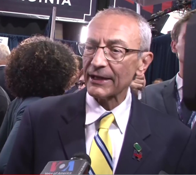 Senator Chuck Schumer in Anzug und Krawatte spricht mit Reportern, während eine Gruppe von Menschen in Mänteln und ID-Karten hinter ihm steht, mit Schildern, die "Sen. Schumer sagt, er sei 'kein Demokrat'".