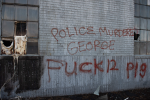 Graffiti an einer Gebäudewand mit der Aufschrift "Polizei ermordet George Fuck 12 P19" mit Fenstern und verstreuten Gegenständen davor