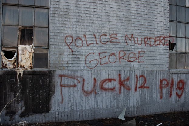 Graffiti an einer Gebäudewand mit der Aufschrift "Polizei ermordet George Fuck 12 P19" mit Fenstern und verstreuten Gegenständen davor