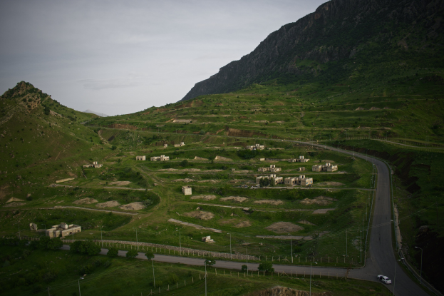 Kleines Dorf in einem Gebirgszug mit grüner Wiese, Bäumen, Pfählen, Fahrzeugen auf einer Straße und sichtbarem Himmel im Hintergrund.