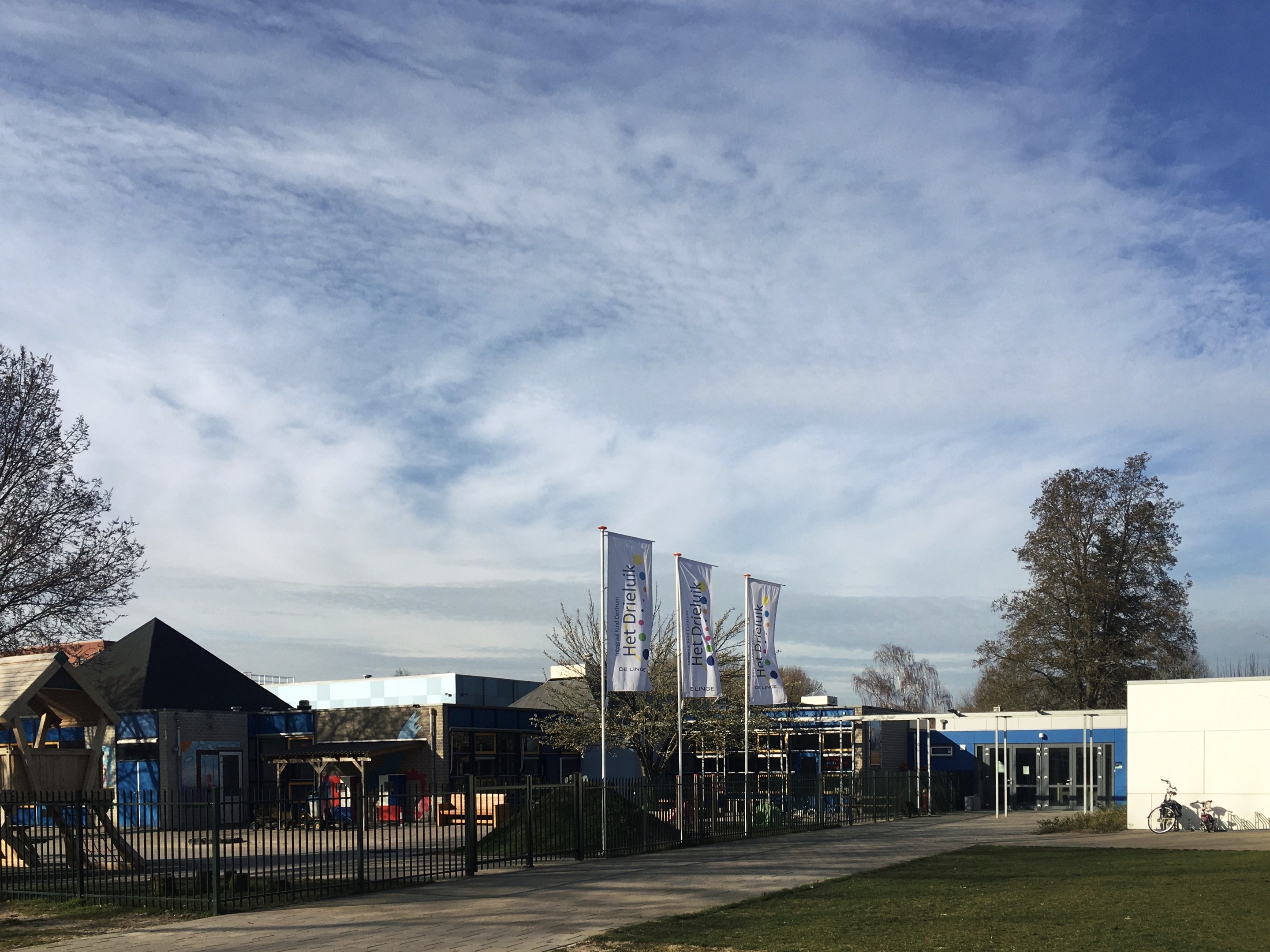 Gro\sses Geb"ude mit einem gr"unen Bereich davor, umgeben von einem Metallzaun, mit einem Weg, B"umen, Pfosten, Bannern, einem geparkten Fahrrad, n"aheren H"usern und einem bew"olkten Himmel, identifiziert als der neue Spielbereich f"ur Kinder in der Schule.