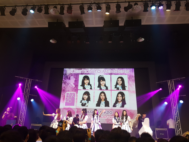 Eine Gruppe von Menschen performs auf einer hell erleuchteten Bühne mit Trägern, während ein Sänger ein Mikrofon hält und ein Musiker ein Instrument spielt, während das Publikum darunter die Veranstaltung aufzeichnet und ein großer Bildschirm Bilder der K-Pop-Gruppe BK48 zeigt.