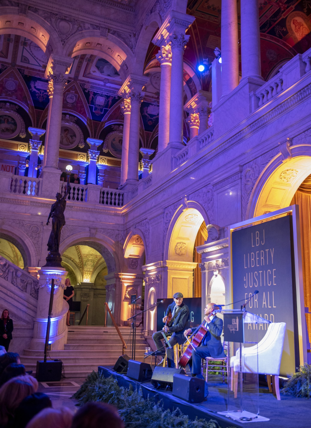 A group of people seated on a stage during the Liberty Justice for All Award Ceremony in New York City, with musicians playing instruments, a podium with a microphone, a board with text, and a statue in the background.