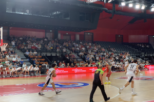 Eine Gruppe von Menschen, die Basketball in einer Turnhalle spielt, mit einem Basketballkorb links und Zuschauern im Hintergrund, unter hellen Stadionlichtern.
