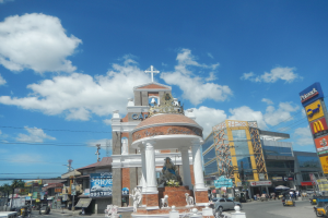 Eine belebte Stadtstraße mit einer Kirche in der Mitte, umgeben von Gebäuden, Geschäften, Schildern, Stromleitungen, Fahrzeugen, Statuen und einem bewölkten Himmel.
