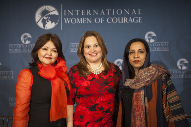 Drei Frauen, die gemeinsam vor einem Banner mit der Aufschrift "Internationale Frauen des Mutes" stehen und lächeln.