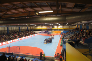 Eine große Halle mit Zuschauern bei einem Handballspiel, mit einem Netz auf der linken Seite, Anzeigetafeln im Hintergrund und Lampen und Rohren an der Decke.