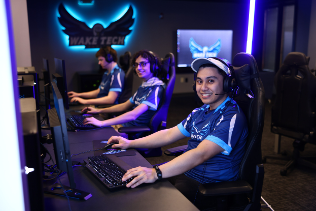 Gruppe von Menschen an Schreibtischen mit Computern, Kopfhörern tragend und lachend, vor Monitoren und Tastaturen, mit einem Logo und Text an der Wand dahinter.