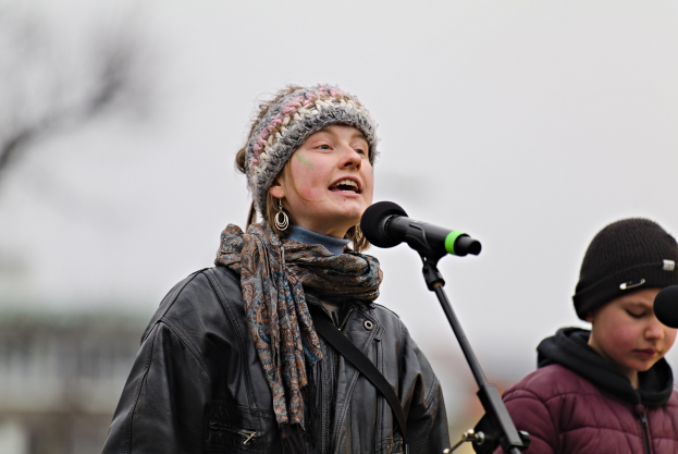 Eine Frau singt in ein Mikrofon auf der Bühne, trägt eine schwarze Jacke, einen Schal und eine Mütze, neben einer anderen Person in einer maronenfarbenen Jacke und schwarzer Mütze vor einem unscharfen Hintergrund.