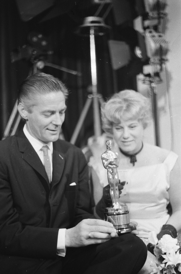 Ein Mann in einem schwarzen Blazer, weißem Hemd und Krawatte hält eine Oscar-Statue und sitzt neben einer Frau, die einen Blumenstrauß hält, während Lichter die schwarze und weiße Szene erhellen.