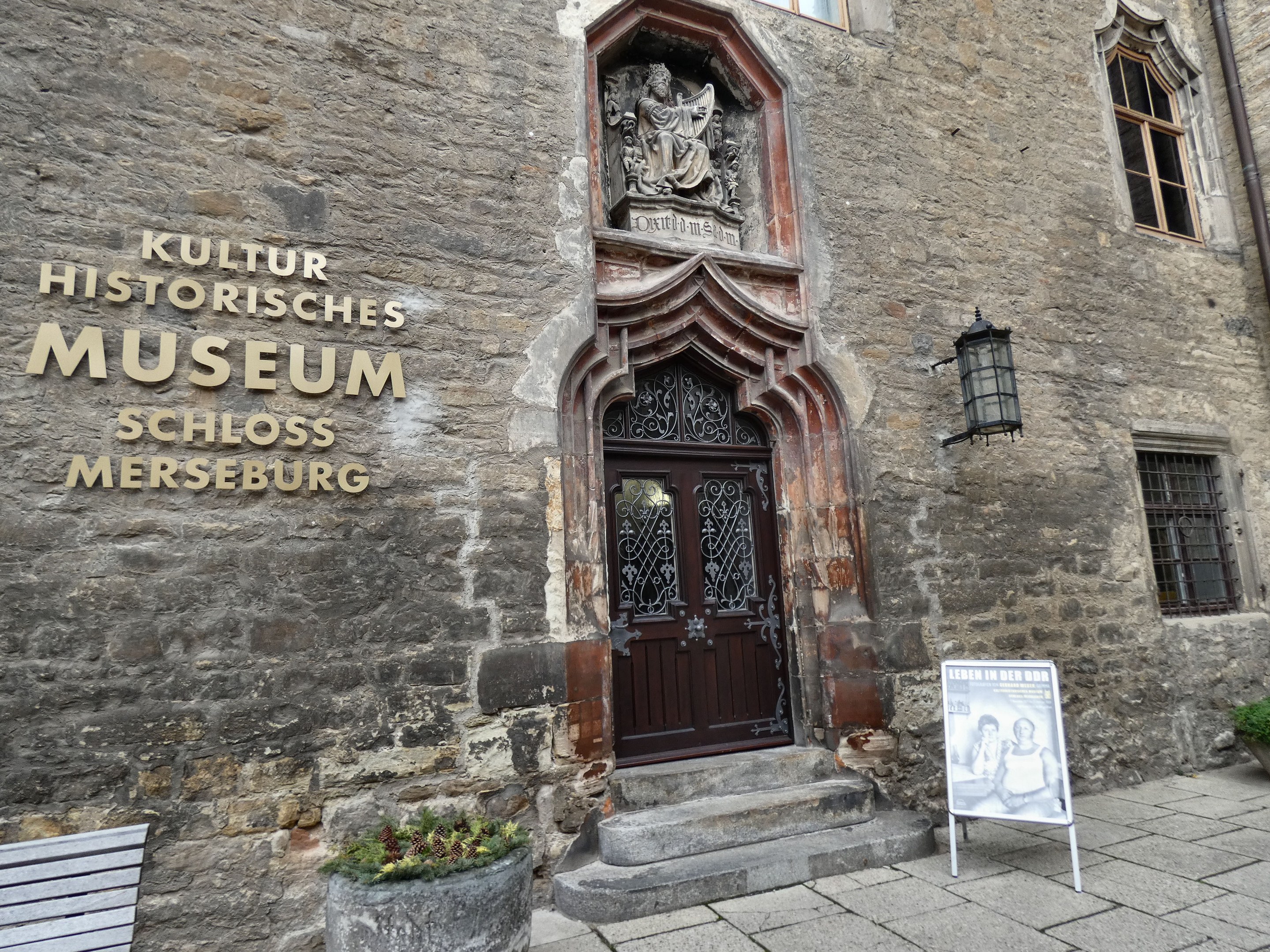 Exterior view of the Kultur Historische Museum in Schloss Merseburg, Germany, featuring windows, a door, a signboard, a bench, potted plants, a pipe, and a cloudy sky.