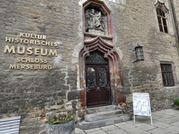 Exterior view of the Kultur Historische Museum in Schloss Merseburg, Germany, featuring windows, a door, a signboard, a bench, potted plants, a pipe, and a cloudy sky.