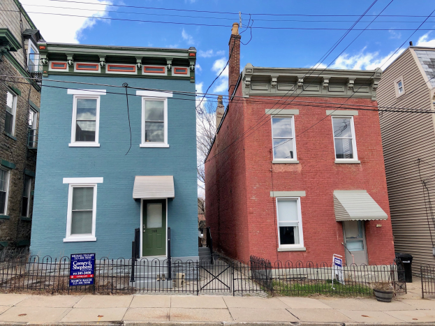 Colorful row of houses with a rental signboard, plants, a trash bin, and a metal fence under a cloudy sky.