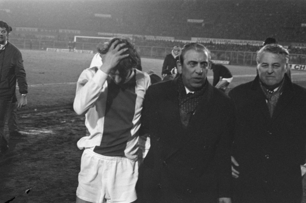 Schwarzes Bild einer Gruppe von Männern auf einem Fußballfeld, die nach einem Spiel enttäuscht aussehen, mit einem Stadion im Hintergrund.