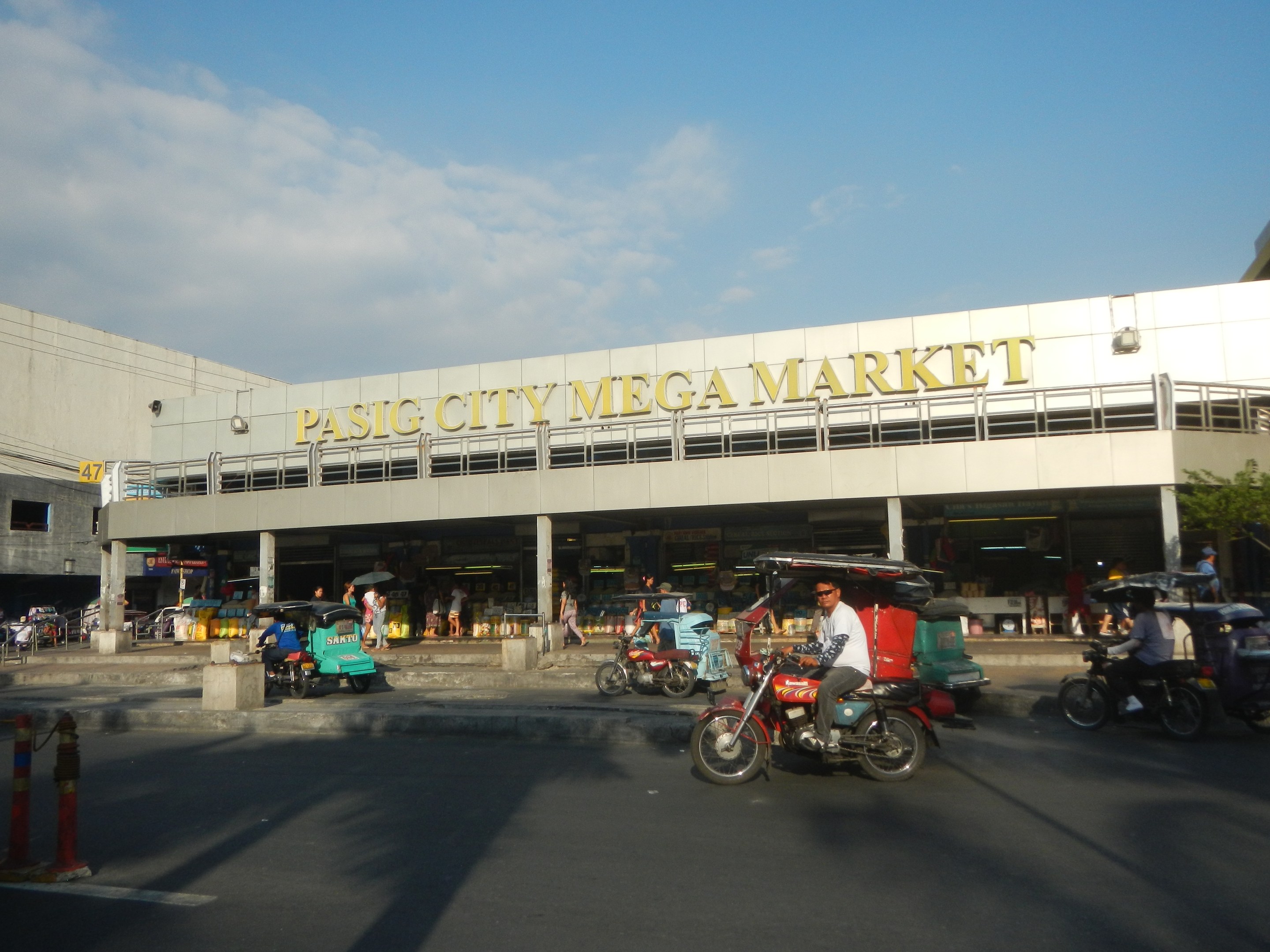 Eine belebte Straßen Szene am Pasig City Mega Market mit Fahrzeugen und Fußgängern, ein Gebäude mit Text im Hintergrund und ein bewölkter Himmel.