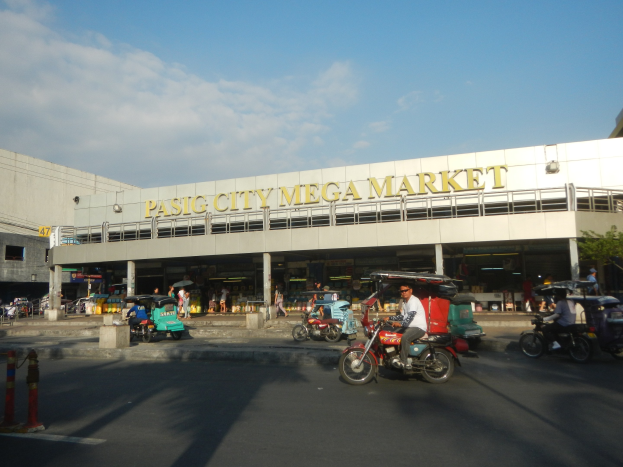 Eine belebte Straßen Szene am Pasig City Mega Market mit Fahrzeugen und Fußgängern, ein Gebäude mit Text im Hintergrund und ein bewölkter Himmel.