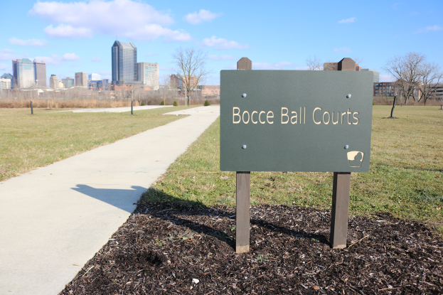 Tafel mit Text "Bocce Ball Courts in Indianapolis, Indiana" auf Gras neben einem Weg, Bäumen, Pfählen, einem Zaun, Gebäuden und einem bewölkten Himmel.